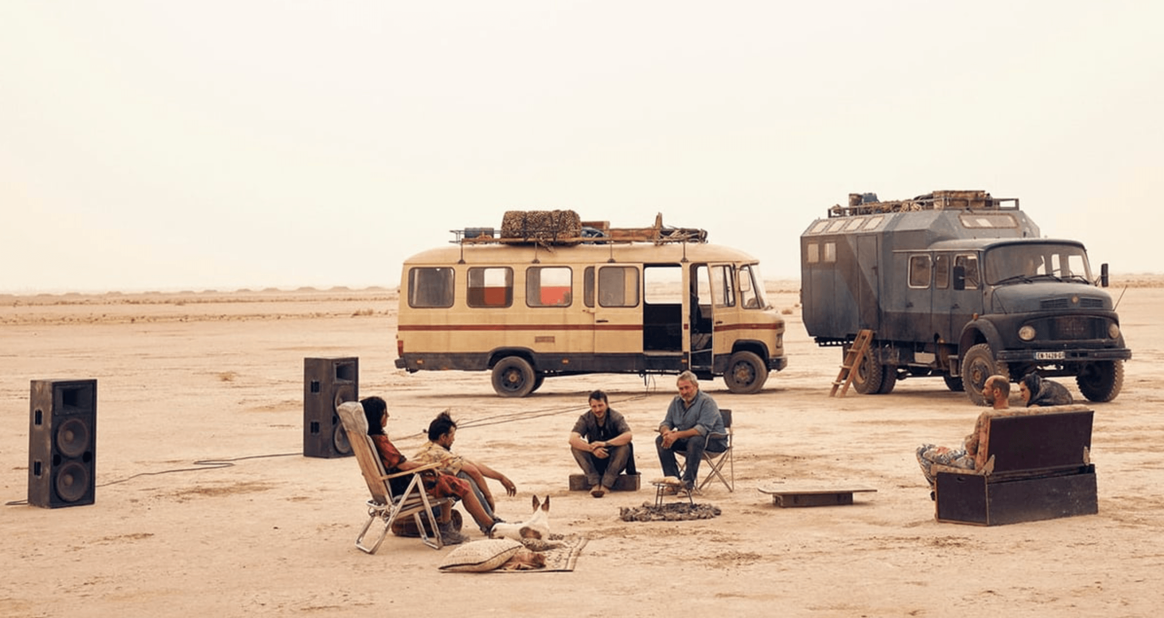 Travelers gather around sound equipment between two vehicles in a desert at dusk in a scene from 'Sirāt'.