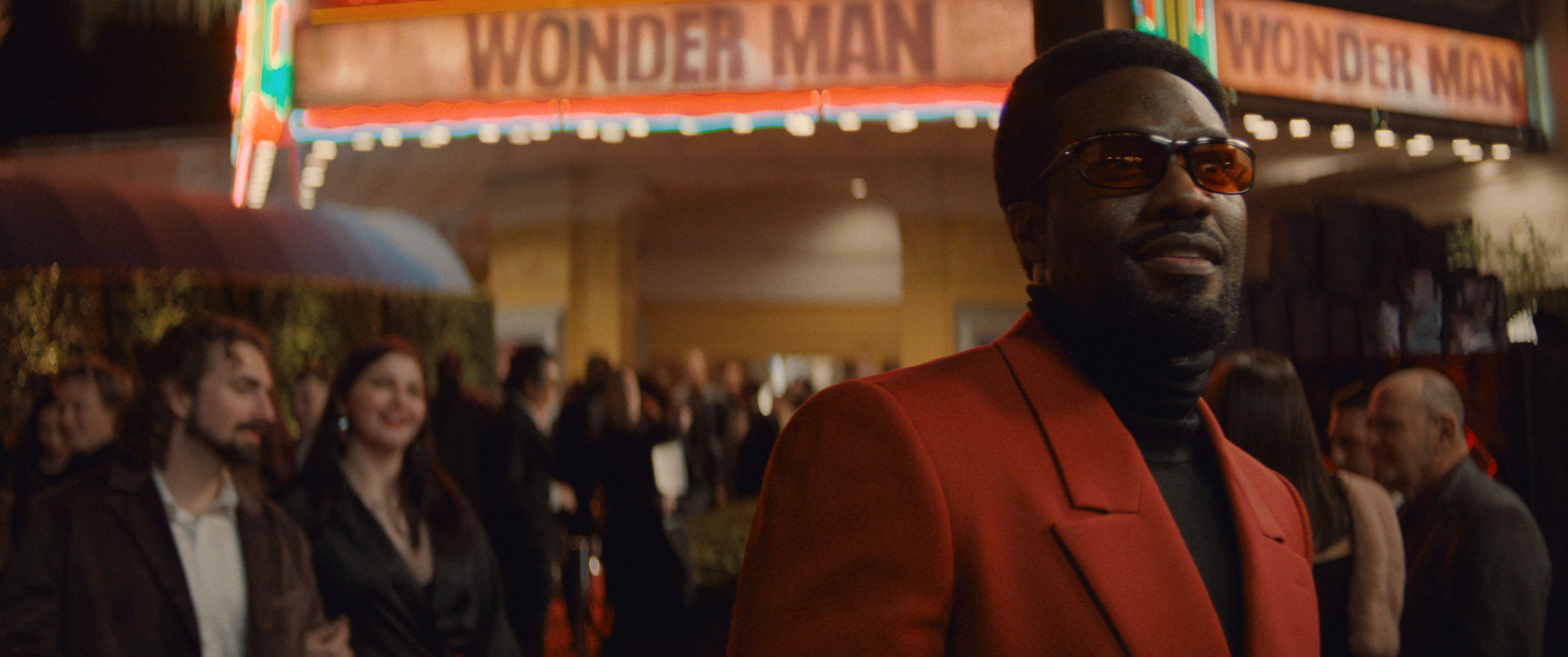 A man in a red blazer and tinted glasses stands under a 'Wonder Man' theater marquee at a premiere event in Marvel's 'Wonder Man'.