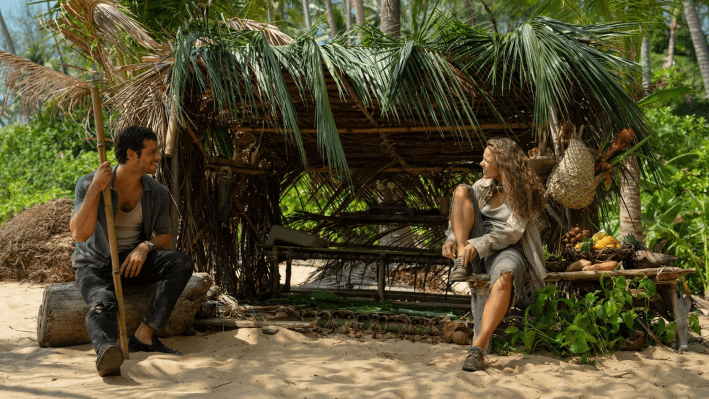 Two castmates sit under a palm-leaf shelter on a remote tropical beach in a scene from 'Send Help'.